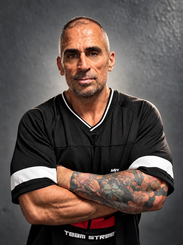 Retrato comercial de un entrenador personal con brazos cruzados sobre fondo gris, sesión de fotografía para Paleo Reset
