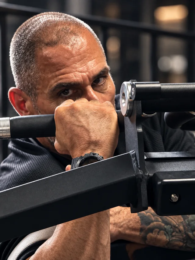 Primer plano de un entrenador personal mirando a cámara entre las barras de una máquina de gimnasio, fotografía de Paleo Reset